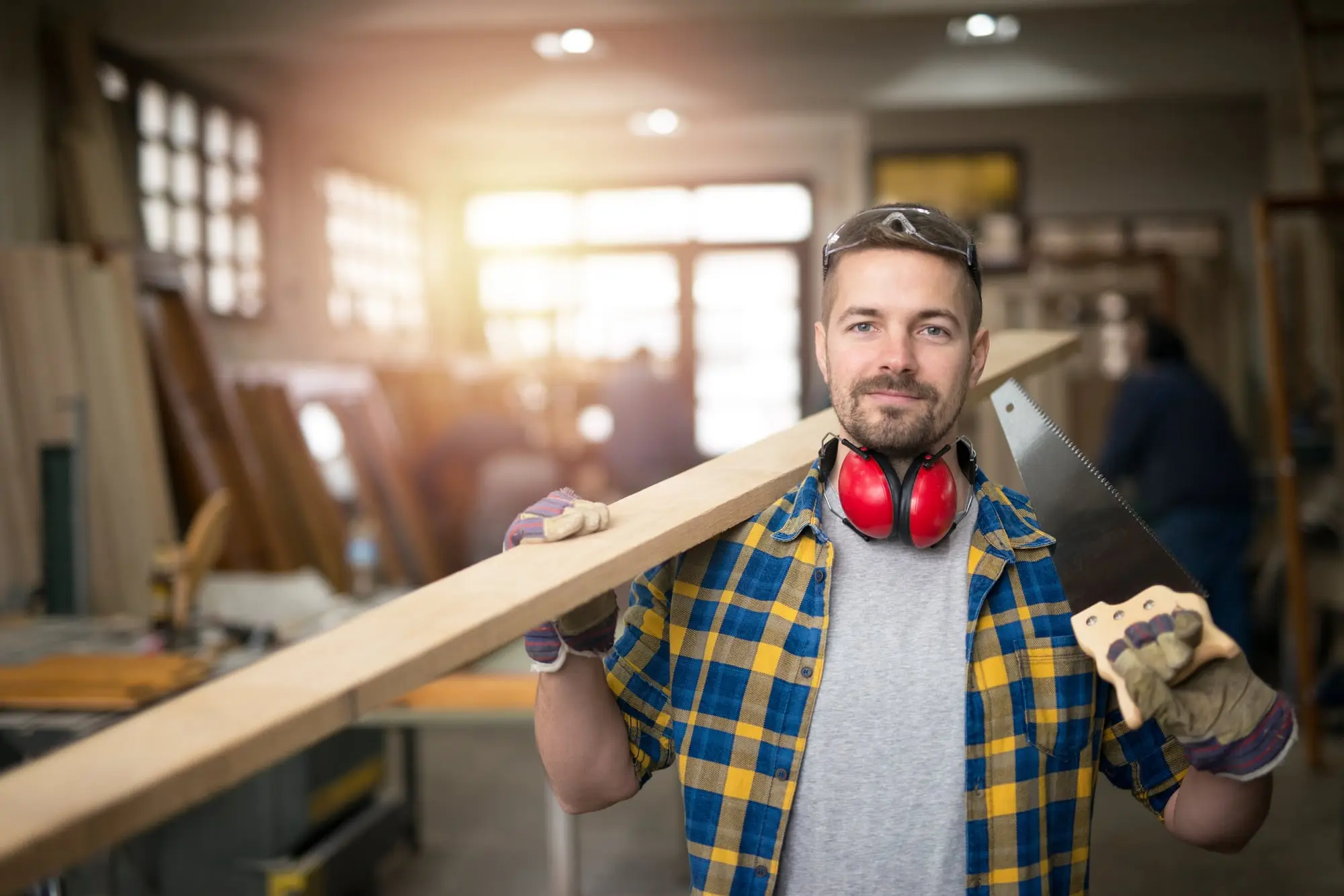 A carpenter in his workshop carrying a plank and a hand saw