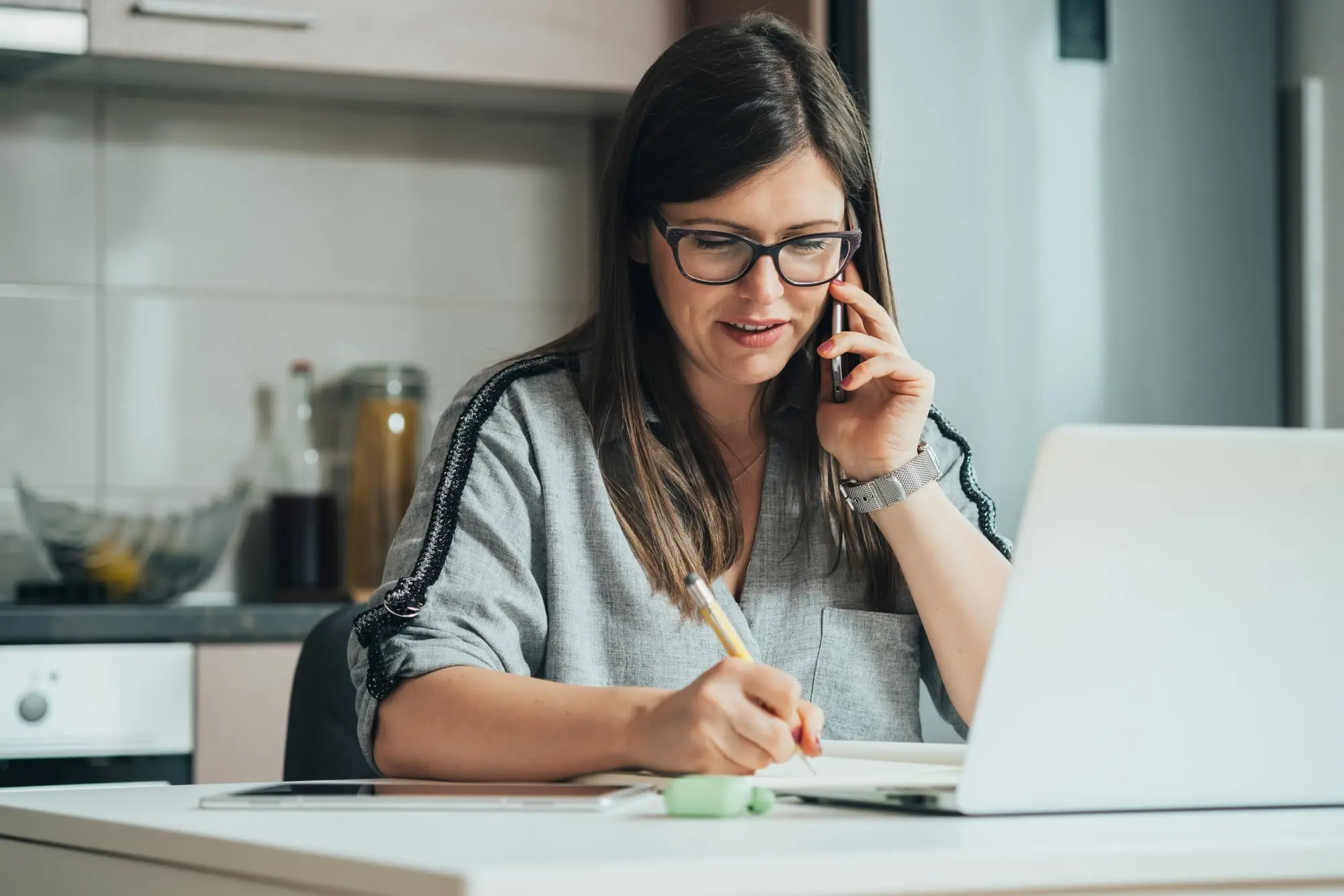A woman on a phone call at her kitchen table, taking notes beside her laptop